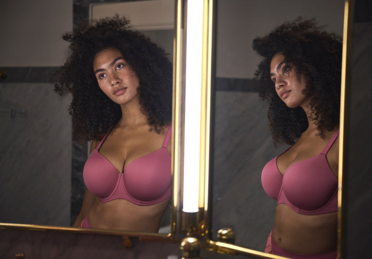 A woman wearing pink bra and panties stands reflected in a vanity mirror